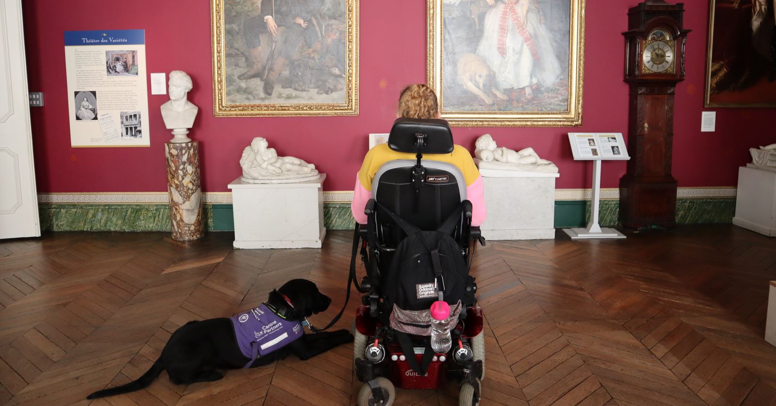 kate Stanforth sat in her wheelchair with support dog looking at paintings within The Bowes Museum